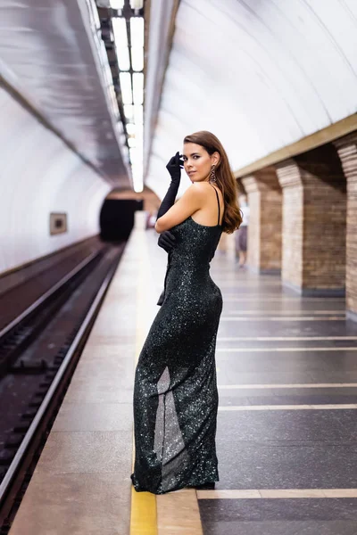 Sinnliche Frau im langen schwarzen Kleid, die auf dem U-Bahnsteig in die Kamera blickt — Stockfoto