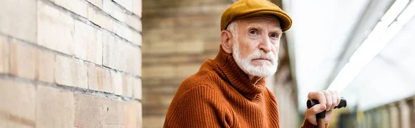 Senior man in sweater and cap looking away while sitting on metro platform on blurred foreground, banner — Stock Photo