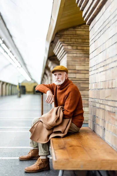 Aged man in autumn outfit leaning on walking stick while sitting on subway platform bench - foto de stock