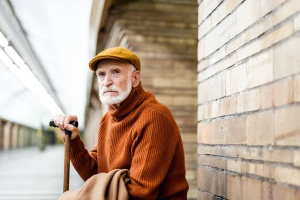 Senior man in sweater and cap looking away while sitting on metro platform with walking stick — Stock Photo