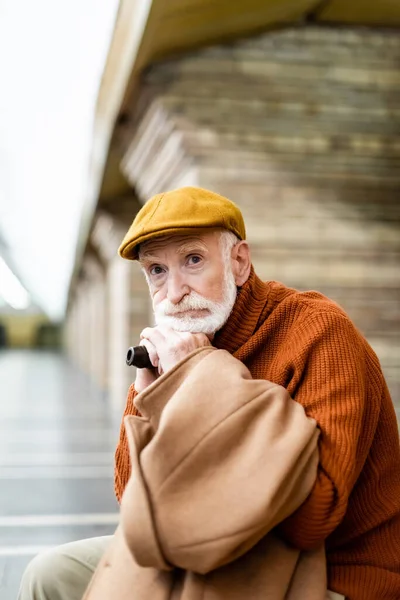 Senior man in cap and sweater looking away while sitting with walking stick on subway platform - foto de stock