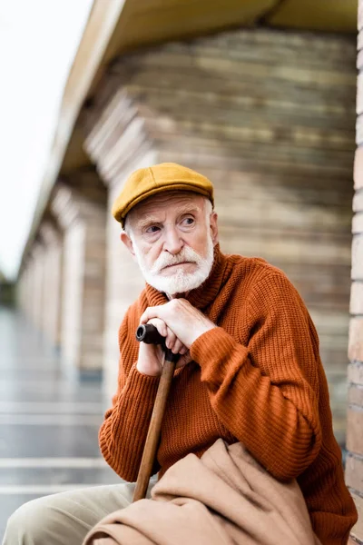 Elderly man in autumn clothes looking away while sitting on underground platform with walking stick — Stock Photo