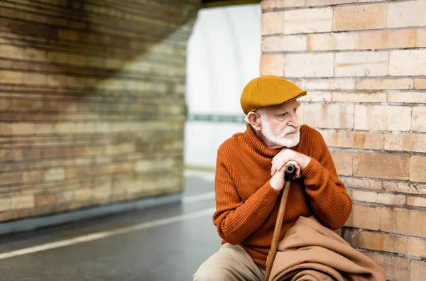 Aged man in cap and sweater looking away while sitting on metro platform bench - foto de stock