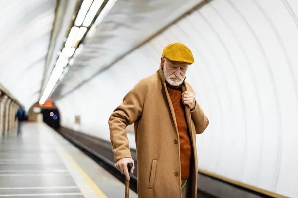 Homme âgé en casquette et manteau d'automne debout avec bâton de marche sur la plate-forme souterraine — Photo de stock