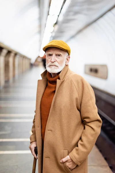 Senior man in autumn cap and coat looking at camera while standing on underground platform — Stock Photo