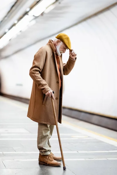 Homme âgé en automne vêtements touchant chapeau tout en se tenant debout avec bâton de marche sur la plate-forme souterraine — Photo de stock