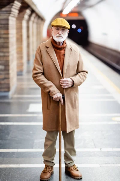 Aged man in autumn outfit looking at camera while standing with walking stick on underground platform - foto de stock