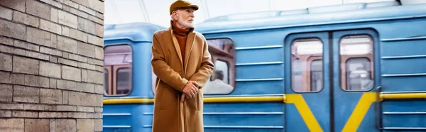Aged man in autumn clothes standing on metro platform with train on background, banner - foto de stock