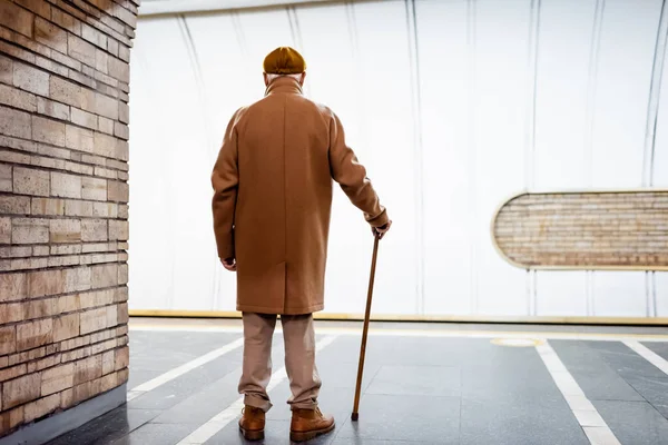 Back view of aged man in autumn outfit standing with walking stick on underground platform — Stock Photo