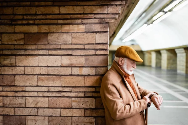 Uomo anziano in abito autunno guardando orologio da polso mentre appoggiato sulla colonna alla stazione della metropolitana — Foto stock