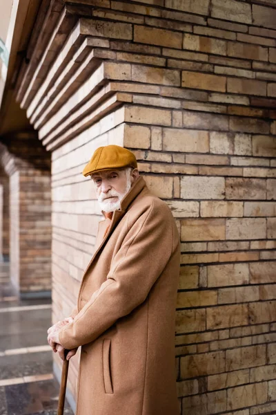 Homme âgé en casquette et manteau d'automne debout avec bâton de marche près de la colonne de brique à la station de métro — Photo de stock