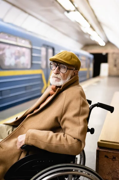 Пожилой инвалид в инвалидной коляске, в осенней одежде, на платформе метро возле размытого поезда — стоковое фото