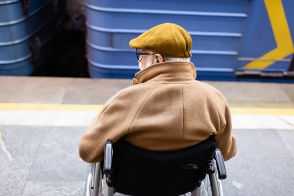 Rückansicht eines älteren behinderten Mannes im Rollstuhl, der Herbstkleidung trägt, in der Nähe eines verschwommenen Zuges auf einer Metrostation — Stockfoto