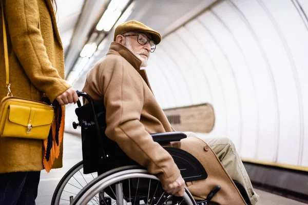 Donna in possesso di sedia a rotelle di anziani handicappati uomo in vestiti autunno sulla stazione della metropolitana — Foto stock