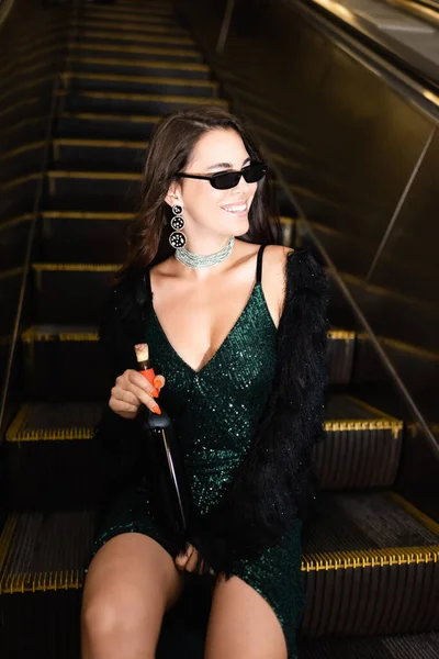 Glamour femme dans des lunettes de soleil assis sur métro escalator avec bouteille de vin — Photo de stock