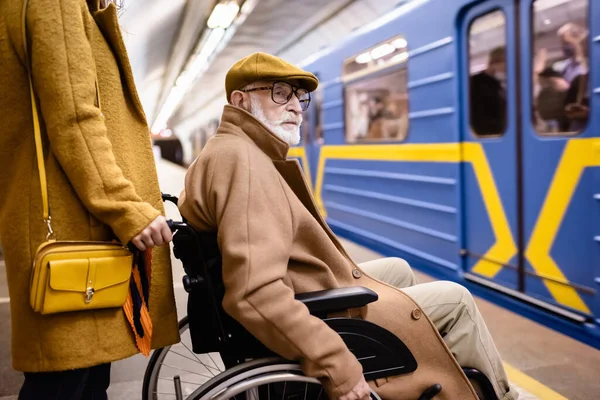 Donna in possesso di sedia a rotelle di un anziano disabile su una piattaforma sotterranea vicino a un treno sfocato — Foto stock