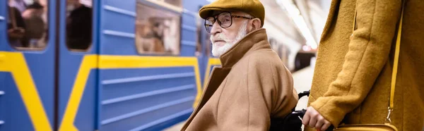 Femme tenant fauteuil roulant d'un homme handicapé âgé sur une plate-forme de métro près d'un train flou, bannière — Photo de stock