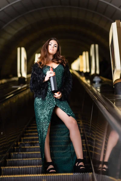 Femme séduisante en robe noire élégante tenant bouteille de vin sur escalator souterrain — Photo de stock