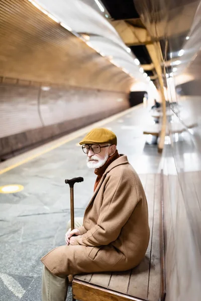 Старший человек в пальто и кепке смотрит в сторону, сидя на подземной скамейке платформы — стоковое фото