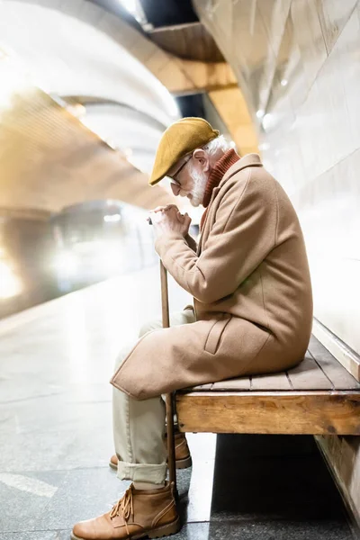 Vista lateral do homem sênior sentado no banco com a cabeça curvada, enquanto o trem que chega na plataforma do metro — Fotografia de Stock