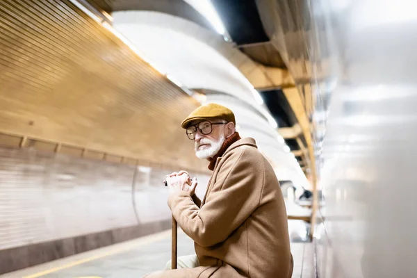 Aged man in autumn outfit and eyeglasses sitting with walking stick on underground platform — Stock Photo