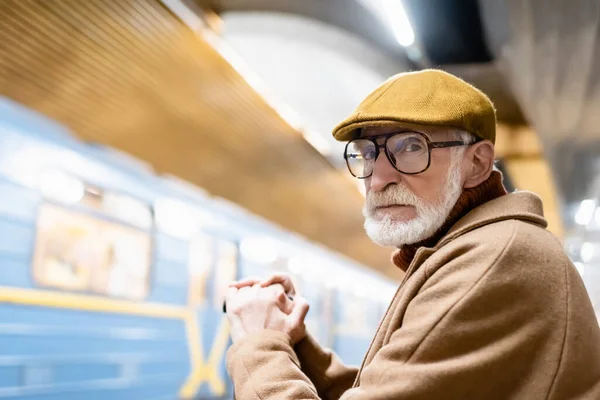 Homem sênior em roupas de outono e óculos olhando para a câmera na plataforma do metrô com trem borrado — Fotografia de Stock