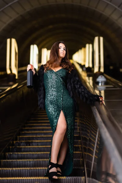 Trendfrau im schwarzen Lurex-Kleid mit einer Flasche Wein auf der Rolltreppe — Stockfoto