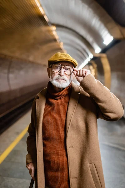 Senior man in autumn cap and coat touching eyeglasses and looking at camera while standing on subway platform — Stock Photo