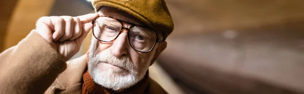 Portrait of aged man in autumn cap touching eyeglasses while looking at camera, banner — Stock Photo