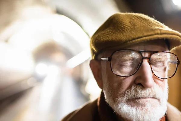 Portrait of aged man in cap and eyeglasses standing with closed eyes on metro platform with light in metro tunnel on background — Stock Photo