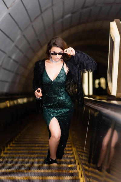 Glamour-Frau im schwarzen Lurex-Kleid berührt Sonnenbrille, während sie auf Rolltreppe in die Kamera schaut — Stockfoto