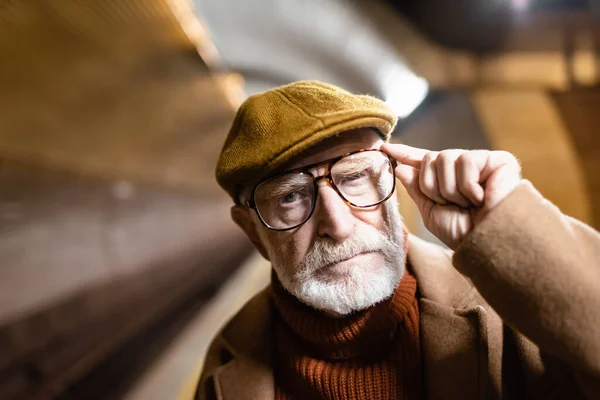 Porträt eines älteren Mannes mit Herbstmütze, der beim Berühren einer Brille in die Kamera blickt — Stockfoto