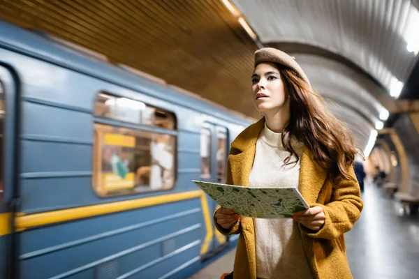 Young woman in beret and autumn coat holding city map near blurred train on subway platform - foto de stock