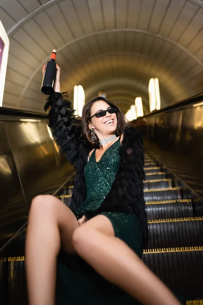 Excited woman in elegant dress holding wine bottle in raised hand while sitting on escalator — Stock Photo