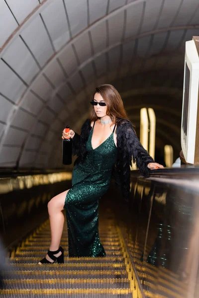 Elegant woman on escalator in long black dress and sunglasses holding wine bottle on blurred foreground — Stock Photo