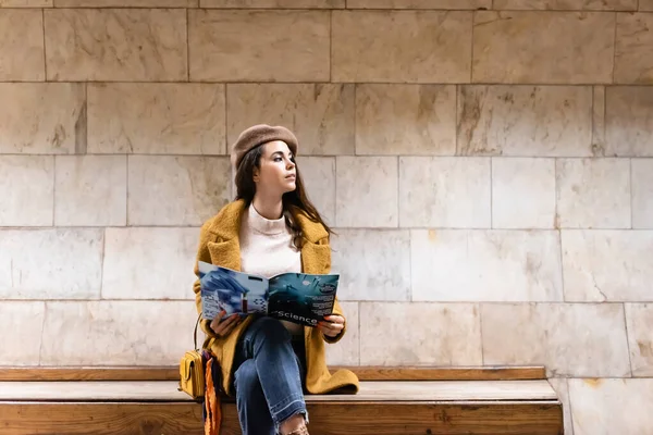 Stylische Frau im Herbst-Outfit schaut weg, während sie Magazin auf U-Bahnsteigbank hält — Stockfoto