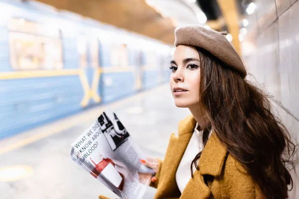 Young woman in beret holding magazine while sitting on metro platform with blurred train — Stock Photo