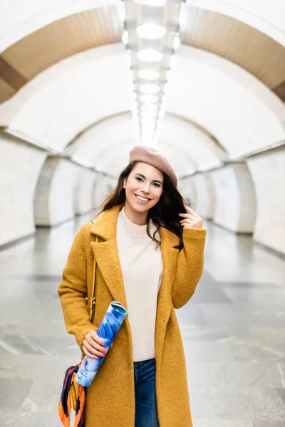 Happy, stylish woman in coat and beret looking at camera while holding magazine at metro station - foto de stock