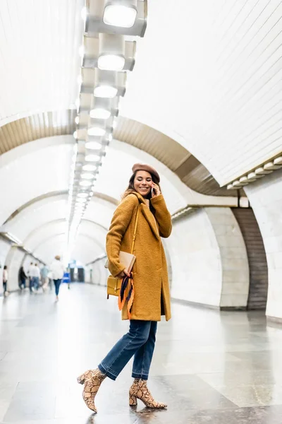 Щаслива жінка в стильному осінньому вбранні розмовляє на смартфоні на підземній станції — стокове фото