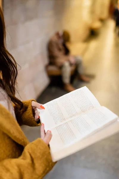 Ausgeschnittene Ansicht einer Frau, die Buch liest, während sie auf dem U-Bahnsteig neben einem alten Mann steht, der auf einer Bank vor verschwommenem Hintergrund schläft — Stockfoto