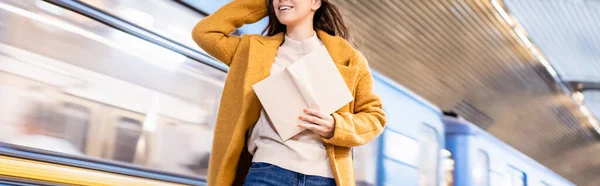 Cropped view of woman in coat holding book while standing on metro platform with blurred train, banner - foto de stock