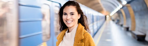 Happy young woman looking at camera on underground platform with blurred train, banner — Stock Photo