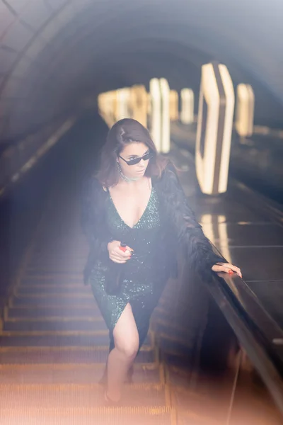 Seductive woman in black dress holding wine bottle and looking away on escalator, blurred foreground - foto de stock