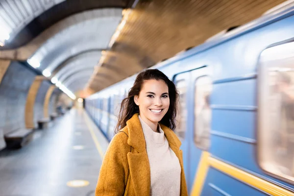 Glückliche Frau im Herbstmantel, die auf dem U-Bahnsteig vor verschwommenem Hintergrund in die Kamera lächelt — Stockfoto