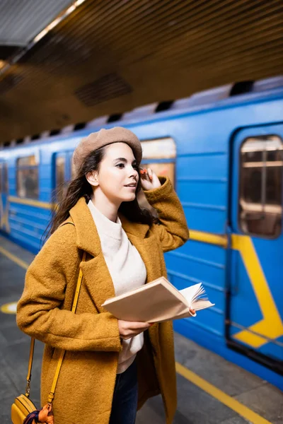 Stylish woman in autumn clothes holding book while looking at blurred train on subway platform - foto de stock