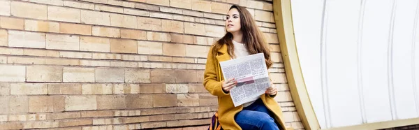Young woman in autumn coat looking away while holding newspaper on subway platform, banner — Stock Photo