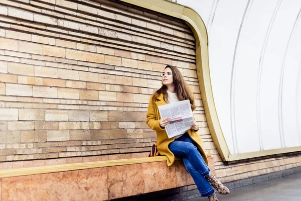 Стильна жінка в осінньому одязі, дивлячись далеко, сидячи на лавці платформи метро з газетою — стокове фото