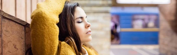 Молодая женщина в осенней одежде стоит с закрытыми глазами и рукой на голове у стены на станции метро, баннер — стоковое фото