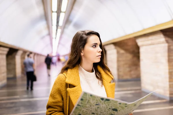 Junge Frau im Herbstmantel schaut weg, während sie am U-Bahnhof einen Stadtplan in der Hand hält — Stockfoto