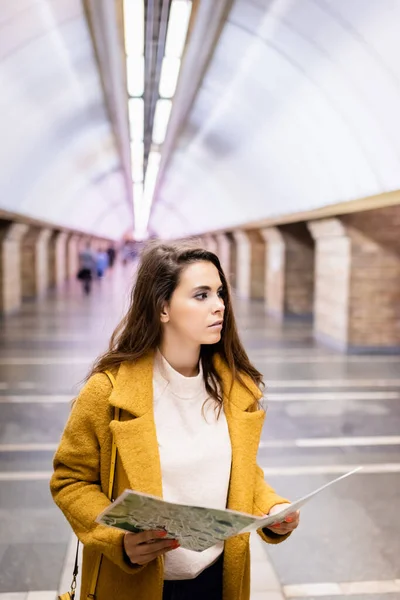 Young woman in stylish autumn coat looking away while holding city map at metro station — Stock Photo